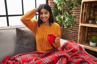 Genç İspanyol kadın kanepede oturmuş deli gibi kahve içiyor ve elleri başının üstünde korkmuş ve ağzı açık bir şekilde şok geçirmekten korkuyor. 