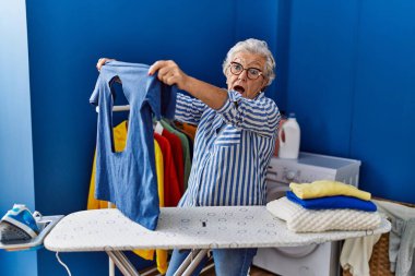 Çamaşırhanede yakılmış demir t-shirt tutan yaşlı, gri saçlı kadın.