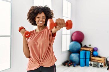 Genç Afrikalı Amerikalı kadın gülümsüyor spor merkezinde halterleri kullanarak kendine güven eğitimi alıyor