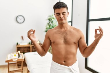 Genç İspanyol adam spa merkezinde üstsüz duruyor rahatlıyor ve gözleri kapalı gülümsüyor parmaklarıyla meditasyon hareketi yapıyor. yoga konsepti. 