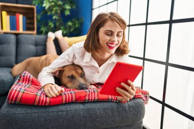 Kafkasyalı genç bir kadın evdeki köpekle birlikte kanepeye uzanmış dokunmatik ped kullanıyor.