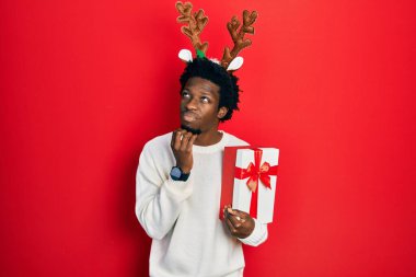 Young african american man wearing deer christmas hat holding gift with hand on chin thinking about question, pensive expression. smiling with thoughtful face. doubt concept. 