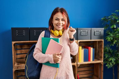Genç beyaz kadın öğrenci çantası ve kitaplarıyla gururla bağırıyor zaferi kutluyor ve başarıyı kollarını kaldırarak kutluyor. 