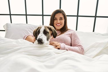Genç kadın gülümsüyor. Kendine güvenen, sarılan köpek yatakta oturuyor.