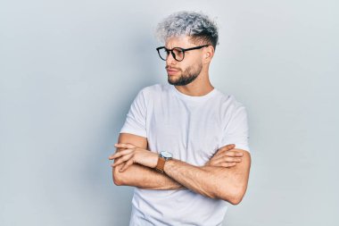 Modern boyalı saçlı, beyaz tişörtlü ve gözlüklü kolları çapraz ve kendinden emin genç İspanyol bir adam. 