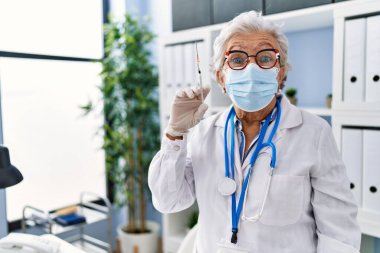 Gri saçlı, doktor üniforması giyen ve elinde şırınga tutan yaşlı bir kadın. Şaşırtıcı bir şekilde ağzı açık kalmış, güvenilmez bir yüz. 