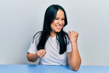 Güzel İspanyol bir kadın, burun piercingli, sıradan beyaz tişörtlü. Masada oturmuş, kolları havada, gözleri kapalı, şaşkınlıkla zaferi kutluyor. 