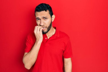 Sakallı, sıradan kırmızı tişörtlü İspanyol bir adam gergin görünüyor ve elleri ağızda tırnak yiyor. anksiyete. 
