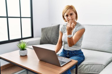 Middle age blonde woman using laptop at home showing middle finger, impolite and rude fuck off expression 