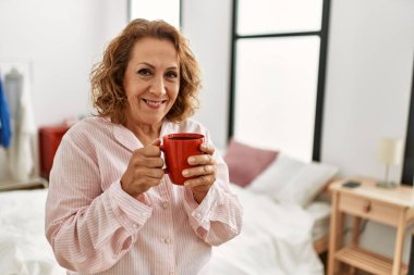 Orta yaşlı beyaz kadın yatak odasında kahve içiyor..