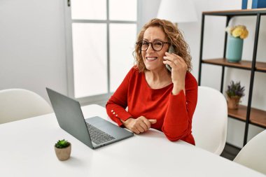 Orta yaşlı beyaz kadın dizüstü bilgisayar kullanıyor ve akıllı telefonla konuşuyor. Evde masada oturuyor..