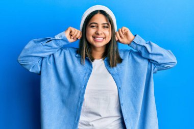 Şirin yün şapkalı, kulakları parmaklarıyla çeken genç Latin bir kadın. Komik bir jest. deneme sorunu 