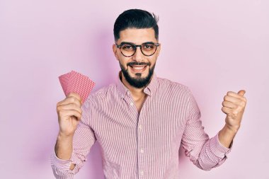 Sakallı, poker kartı tutan yakışıklı bir adam başparmağını kaldırmış, ağzı açık gülümsüyor.  