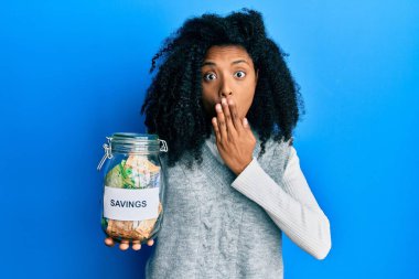 Afro saçlı Afrikalı bir kadın, elinde tasarruf kavanozuyla Güney Afrika 'dan gelen parayla ağzı kapalı, şok olmuş ve hata yapmaktan korkan bir kadın. şaşırmış ifade 