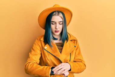 Sarı şapkalı ve deri ceketli genç modern bir kız kol saatini kontrol ediyor, rahat ve kendinden emin. 