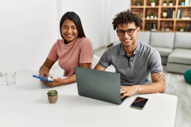 Genç latin çift evde masanın üzerinde dizüstü bilgisayar ve dokunmatik ped kullanarak çalışıyor..