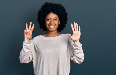 Genç Afro-Amerikalı kadın günlük giysiler giyiyor ve 8 numaralı parmaklarıyla işaret ederken kendinden emin ve mutlu gülümsüyor.. 