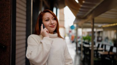 Genç kızıl saçlı, gülümseyen, kendine güvenen, caddedeki akıllı telefondan konuşan kadın.