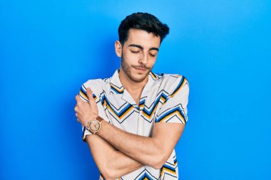 Gündelik kıyafetler giyen genç İspanyol adam kendine sarılıyor mutlu ve pozitif, kendinden emin gülümsüyor. Sevgi ve özgüven. 