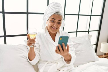 Orta yaşlı, kır saçlı, şampanya içen ve yatak odasında akıllı telefon kullanan bir kadın..