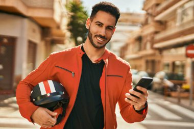 Genç İspanyol adam şehirde moto miğferi tutan akıllı telefon kullanıyor..