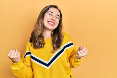 Genç İspanyol kız gündelik kıyafetler giyiyor. Çok mutlu ve heyecanlı. Kazanan jest yapıyor, kollarını kaldırıyor, gülümsüyor ve başarı için bağırıyor. kutlama konsepti. 