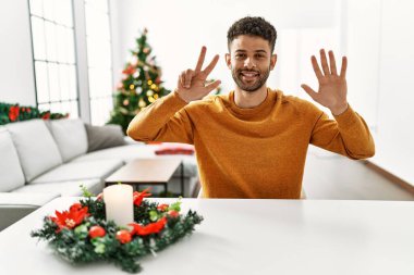 Arap genç adam Noel ağacının yanında oturuyor ve 8 numaralı parmaklarıyla işaret ediyor kendinden emin ve mutlu bir şekilde gülümsüyor.. 