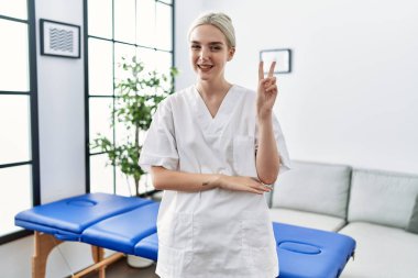 Genç Kafkas fizyoterapist kadın evde çalışıyor. Gülümsüyor, kameraya göz kırpıyor ve zafer işareti yapıyor. İki numara.. 