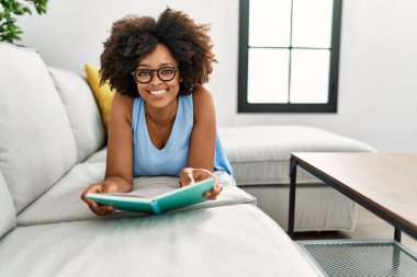 Genç Afro-Amerikan kadın gülümseyerek evde kendinden emin bir şekilde kitap okuyor.