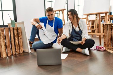 Resim yaparken gülümseyen iki öğrenci sanat okulunda yerde oturuyor..