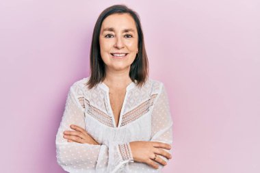 Orta yaşlı İspanyol kadın gündelik kıyafetler giyip kollarını kavuşturup kameraya bakarken gülümsüyor. pozitif kişi. 