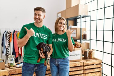 Bağışta gönüllü tişört giyen tatlı köpekli genç çift baş parmağıyla yan tarafa bakıyor ve ağzı açık gülümsüyor. 