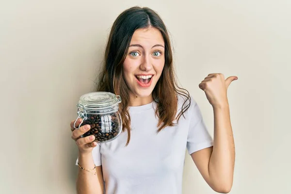 Young Brunette Woman Holding Jar Coffee Beans Pointing Thumb Side — стоковое фото