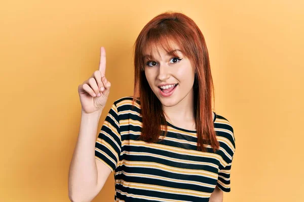 红头发的年轻女性穿着休闲的条纹T恤 面带微笑 带着一个想法或问题指指点点 面带笑容 位居第一 — 图库照片