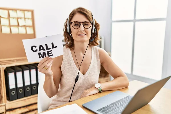 Middle age brunette woman wearing operator headset holding call me banner winking looking at the camera with sexy expression, cheerful and happy face.
