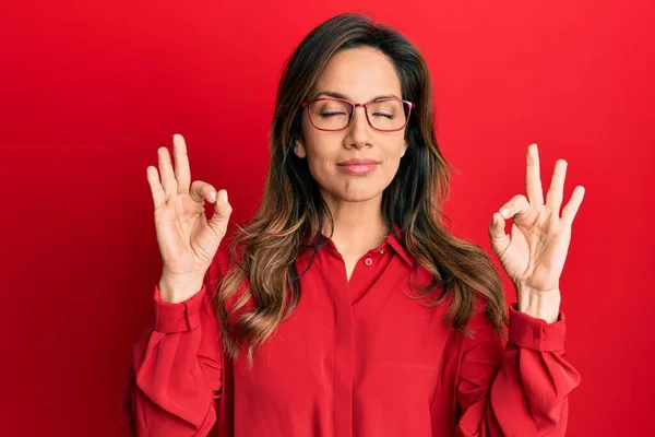 カジュアルな服と眼鏡を身に着けている若いラテン語の女性は 指で瞑想ジェスチャーを行う閉じて目でリラックスして笑顔 ヨガのコンセプト — ストック写真