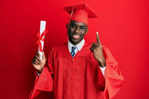 卒業キャップと卒業式のローブを身に着けている若いアフリカ系アメリカ人男性幸せな顔をしたアイデアや質問指差し指で笑顔卒業証書を保持 ナンバーワン — ストック写真