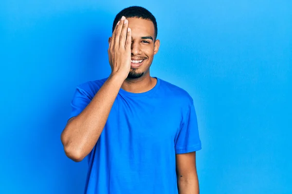 若いアフリカ系アメリカ人の男性が青いシャツを着て 片手で片目を覆い 顔に自信を持って笑顔と驚きの感情 — ストック写真