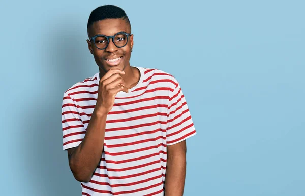 カジュアルなTシャツを着た若いアフリカ系アメリカ人の男性と カメラに自信を持っている眼鏡をかけ 両腕を交差させ 顎に手を上げた ポジティブな — ストック写真