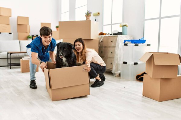 Молодая кавказская пара улыбается счастливой транспортировке собаки в картонную коробку в качестве автомобиля в новом доме.