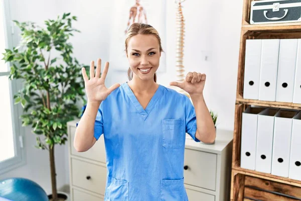 Young caucasian woman working at pain recovery clinic showing and pointing up with fingers number six while smiling confident and happy.