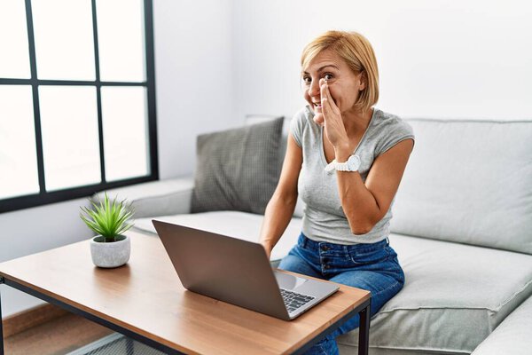 Блондинка средних лет, использующая ноутбук дома, рука об руку рассказывает тайные слухи, шепчет злобные разговоры 