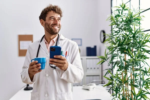 Joven Hispano Vistiendo Uniforme Médico Usando Smartphone Tomando Café Clínica —  Fotos de Stock