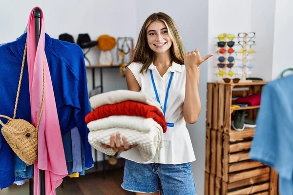 Młoda Blondynka Pracuje Jako Menedżer Detalicznym Butiku Trzymając Swetry Wskazując — Zdjęcie stockowe