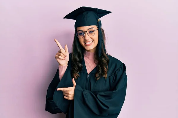若いですヒスパニック系の女性は卒業キャップと式のローブを身に着けています笑顔とカメラを見て2本の手と指で側面を指して — ストック写真