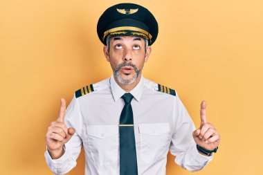 Gri saçlı, uçak pilotu üniforması giyen yakışıklı orta yaşlı bir adam şaşırmış ve şaşırmış bir şekilde yukarı bakıyor ve parmaklarıyla işaret ediyor ve kollarını kaldırıyordu.. 
