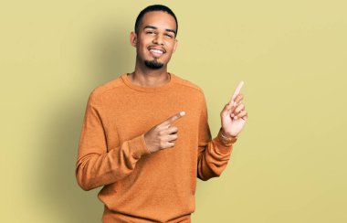 Genç Afro-Amerikalı sıradan kıyafetler giyen, gülümseyen ve kameraya bakan iki elini ve parmağını yana doğru gösteren bir adam.. 