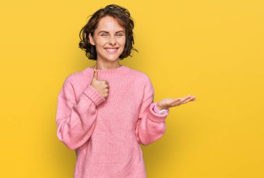 Latin asıllı genç bir kadın, günlük kıyafetler giyiyor, avuç içi gösteriyor ve baş parmaklarıyla iyi bir jest yapıyor, mutlu ve neşeli gülümsüyor. 