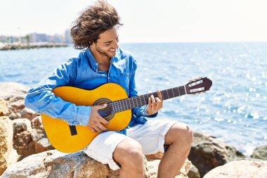 Genç İspanyol adam klasik gitar çalıyor plajda rock 'ın üzerinde oturuyor..