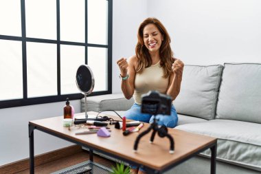 Orta yaşlı İspanyol bir kadın evinde akıllı telefonuyla makyaj yapıyor çok mutlu ve heyecanlı bir şekilde kollarını kaldırarak, gülümseyerek ve başarı çığlıkları atarak zafer hareketi yapıyor. kutlama konsepti. 